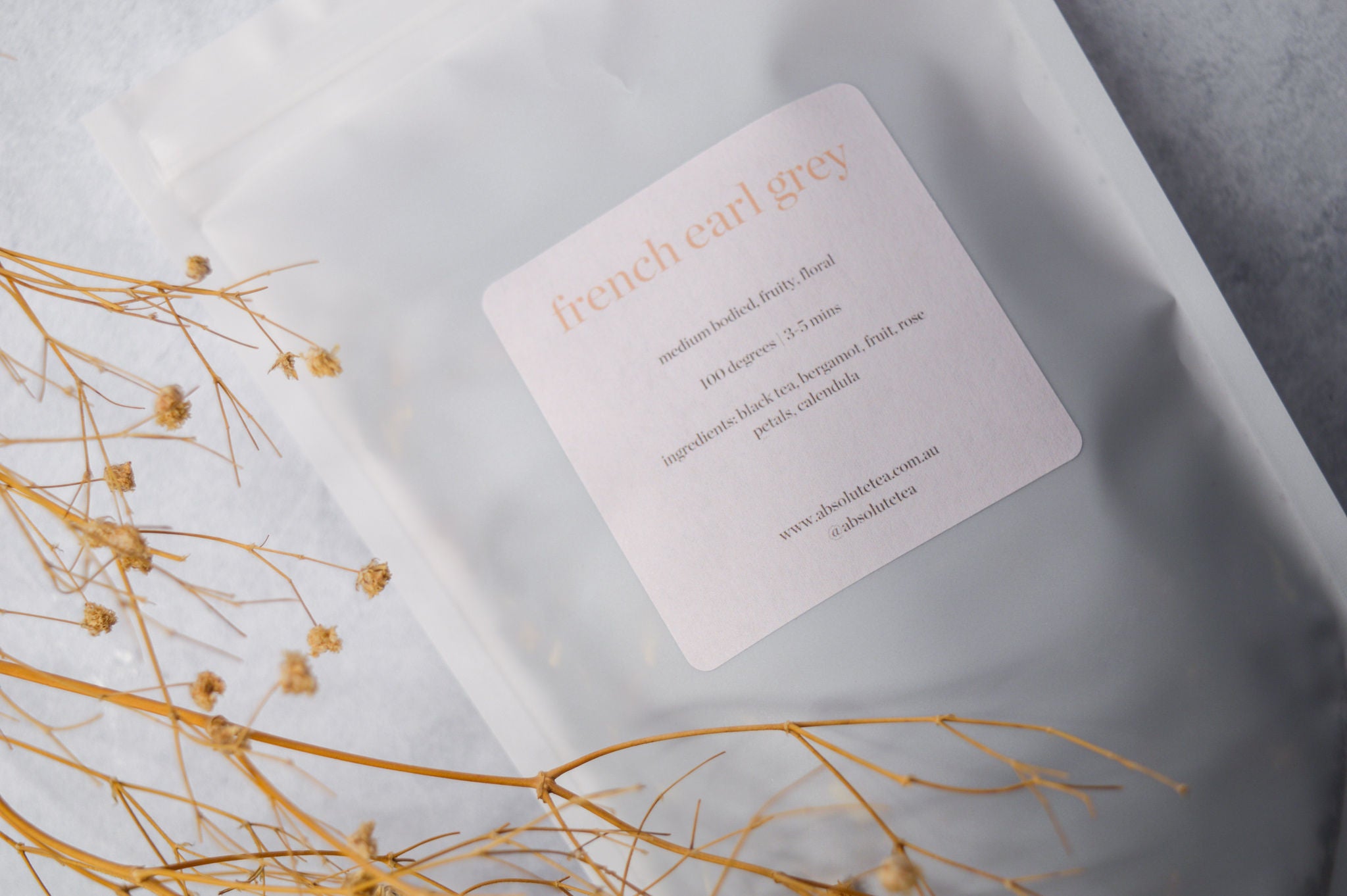 a white translucent zip seal (recyclable) pouch lay filled with French Earl Grey loose leaf tea. A dried stem of baby's breath sits in the foreground. The label on the pouch of tea contains the blend, the ingredients and how to brew. 